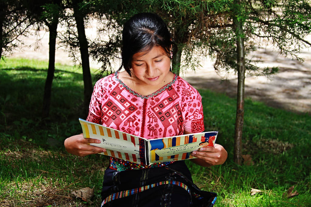 Little girl wearing a pin and read shirt sits in the grass and reads her workbook while grinning.